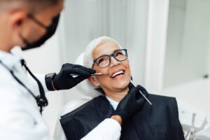 A smiling older woman with short gray hair and glasses sits in a dental chair while a dentist in gloves and a mask examines her teeth, much like an expert reviewing data on an SEO pipeline.