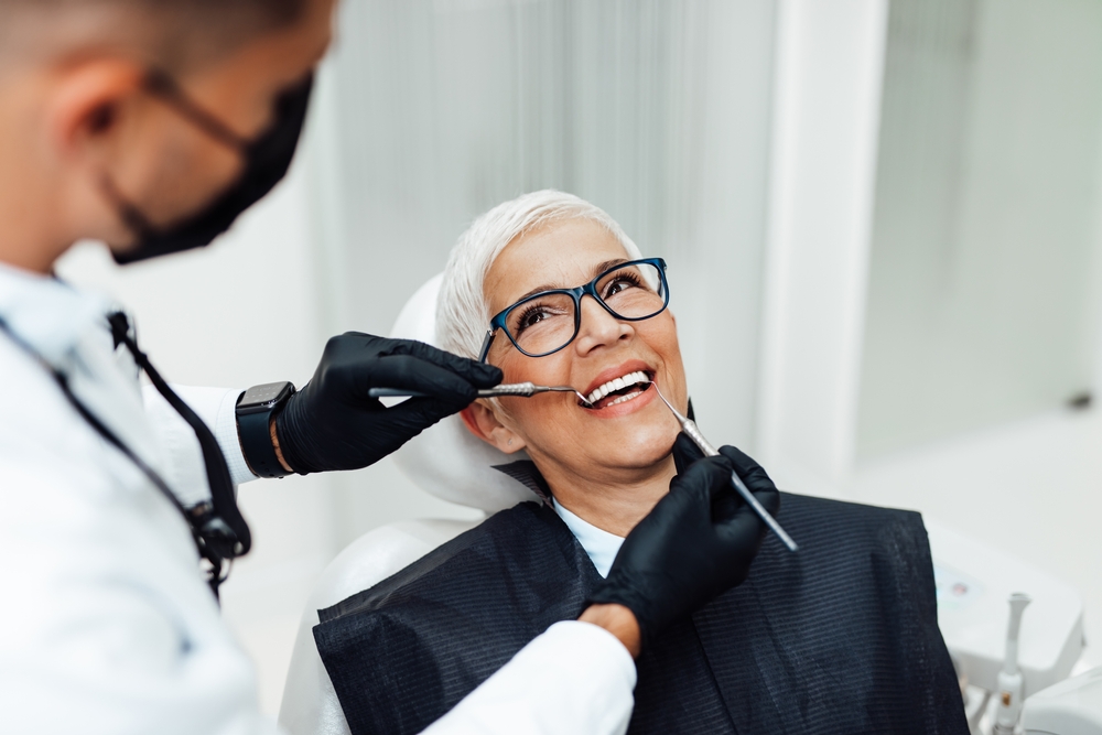 A smiling older woman with short gray hair and glasses sits in a dental chair while a dentist in gloves and a mask examines her teeth, much like an expert reviewing data on an SEO pipeline.